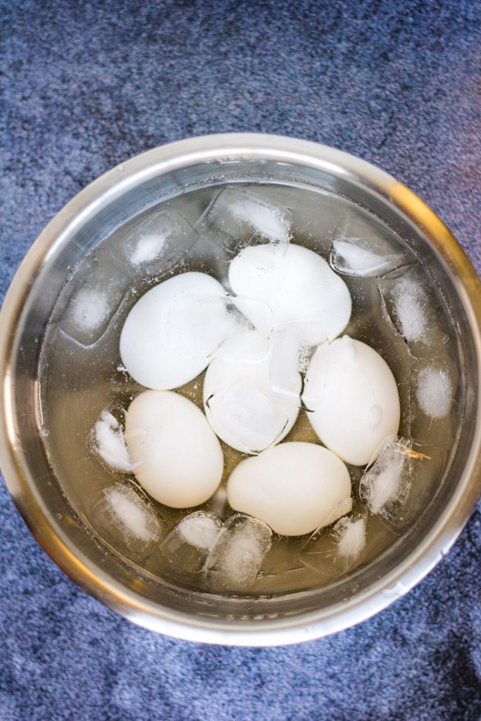 A stainless steel bowl containing white eggs and ice cubes in water, placed on a blue surface.