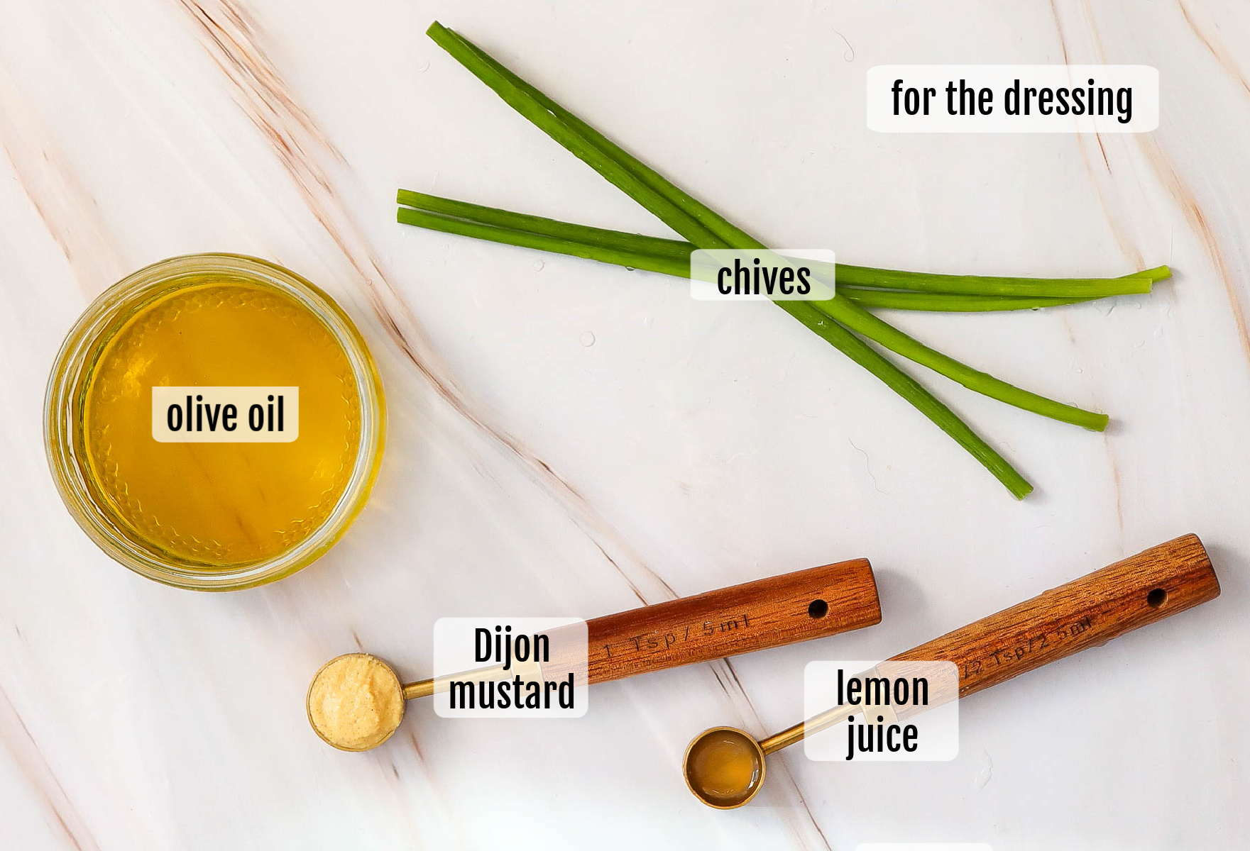 A glass jar of olive oil, three chive stalks, a wooden spoon of Dijon mustard, lemon juice, and labels indicating ingredients for the dressing—perfect for drizzling over asparagus or tossing into a vibrant pea salad.