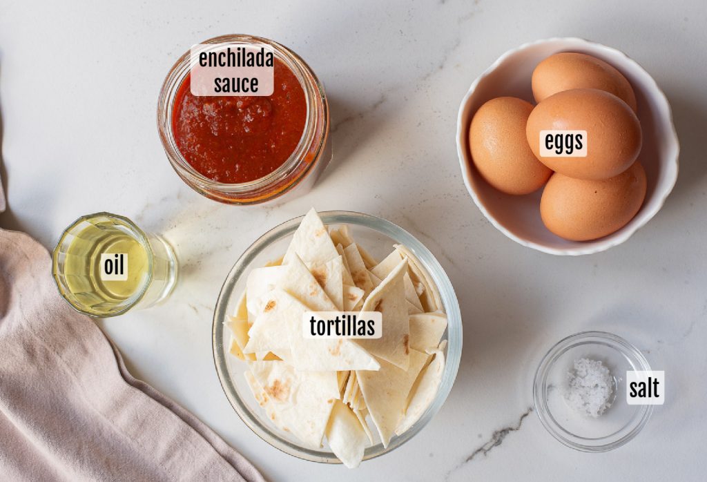 Bowls containing enchilada sauce, eggs, cut tortillas, oil, and salt are arranged on a white countertop for this Chilaquiles Rojos recipe, with each ingredient labeled—perfect for making authentic Chilaquiles Rojos with Fried Eggs.