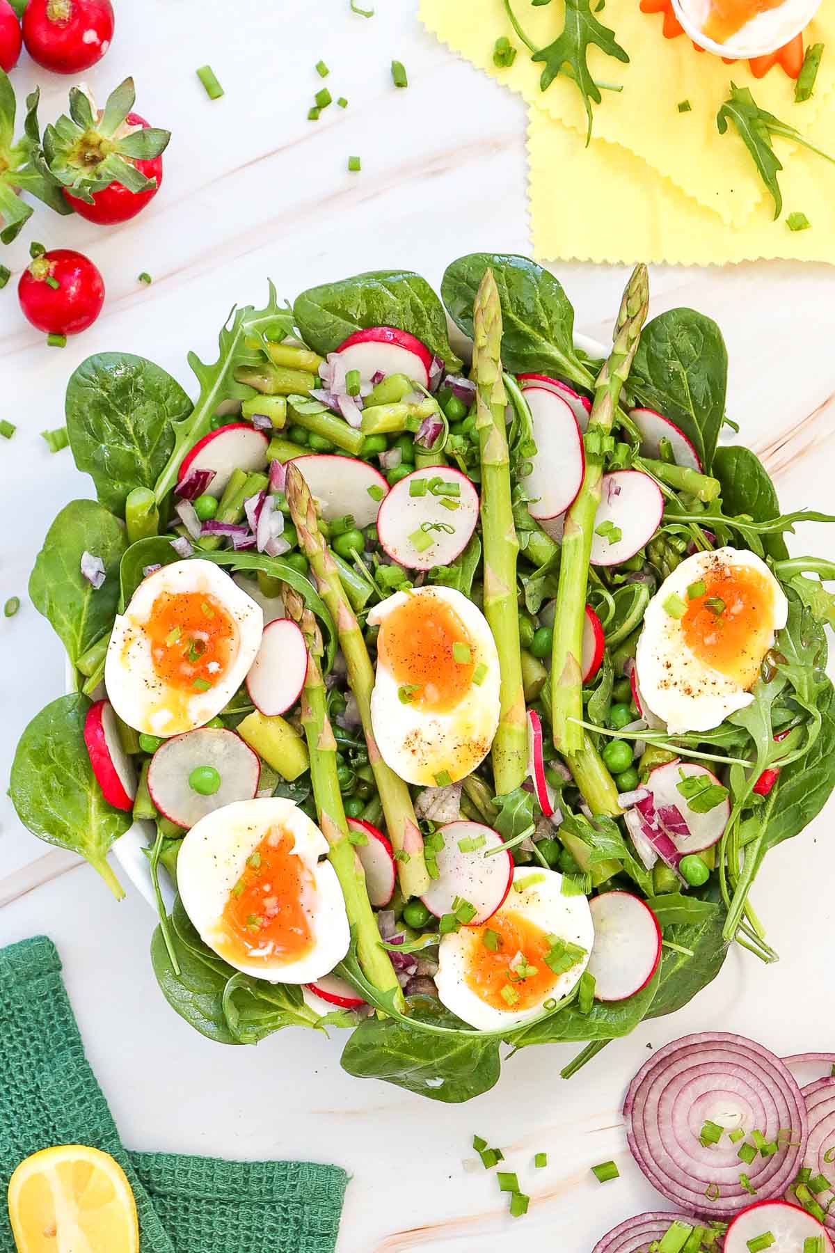 A vibrant pea salad with spinach, asparagus, radishes, red onions, green beans, and soft-boiled eggs topped with orange sauce is arranged on a white plate.