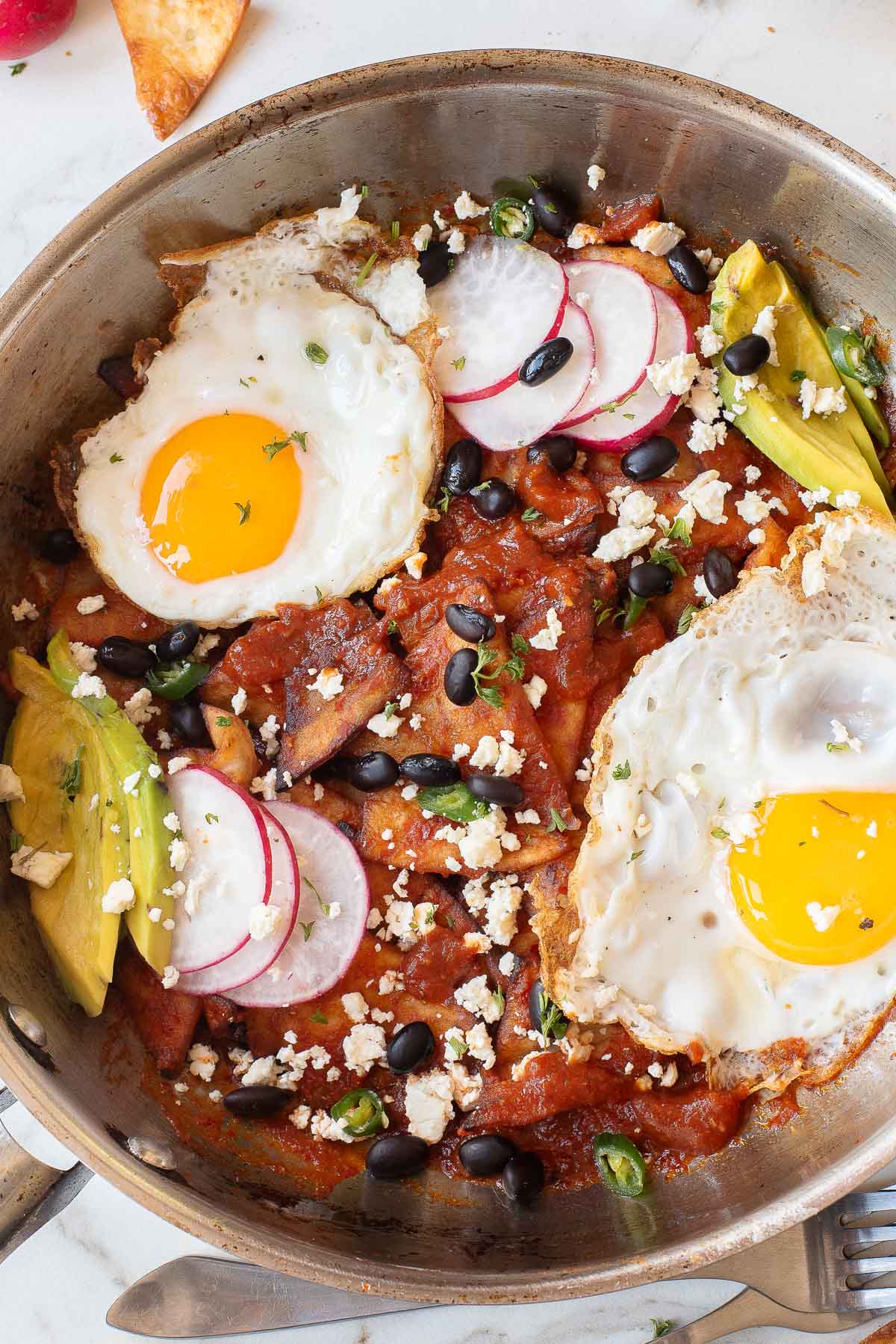 A skillet with Chilaquiles Rojos topped with fried eggs, sliced radishes, avocado, black beans, cheese, and green peppers. This delicious Chilaquiles Rojos recipe is perfect for a hearty breakfast or brunch.