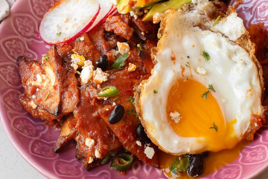 A plate of Chilaquiles Rojos topped with black beans, sliced radishes, green onions, crumbled cheese, and a perfectly cooked fried egg with a runny yolk.