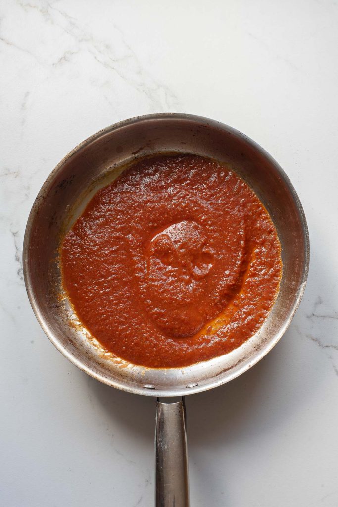 A stainless steel pan containing a layer of red tomato sauce for Chilaquiles Rojos sits on a white marble countertop.
