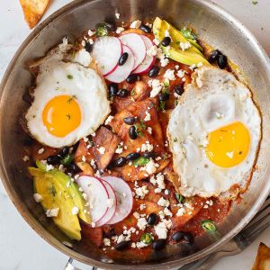 A skillet with Chilaquiles Rojos topped with fried eggs, sliced avocado, radishes, black olives, and crumbled cheese.