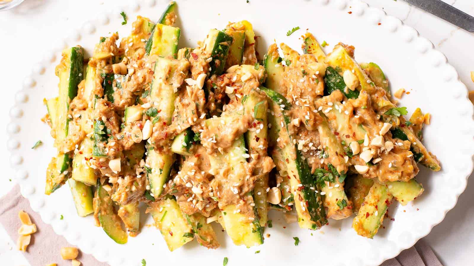 A white oval plate with cucumber spears topped with a creamy peanut sauce, chopped peanuts, herbs, and red pepper flakes.