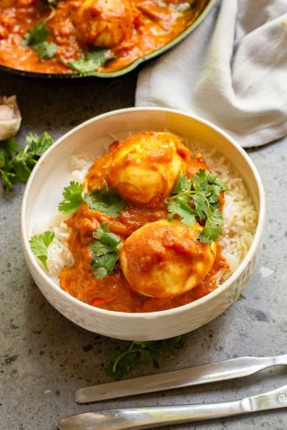 A bowl of white rice topped with two hard-boiled eggs in a rich Indian-Style Egg Curry tomato sauce, garnished with cilantro. A fork and knife are beside the bowl.