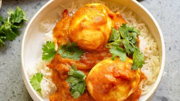 A bowl of white rice topped with two boiled eggs in a vibrant Indian-Style Egg Curry sauce, garnished with fresh cilantro leaves.