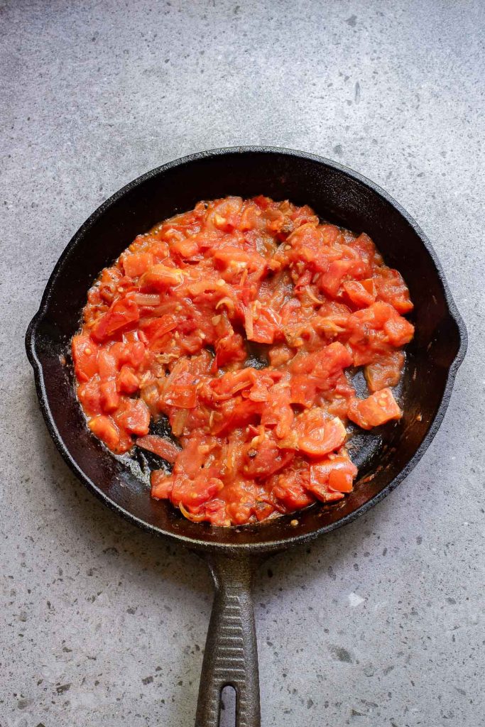 Chopped tomatoes cooking in a black cast iron skillet on a gray countertop, perfect for starting an Indian-Style Egg Curry.