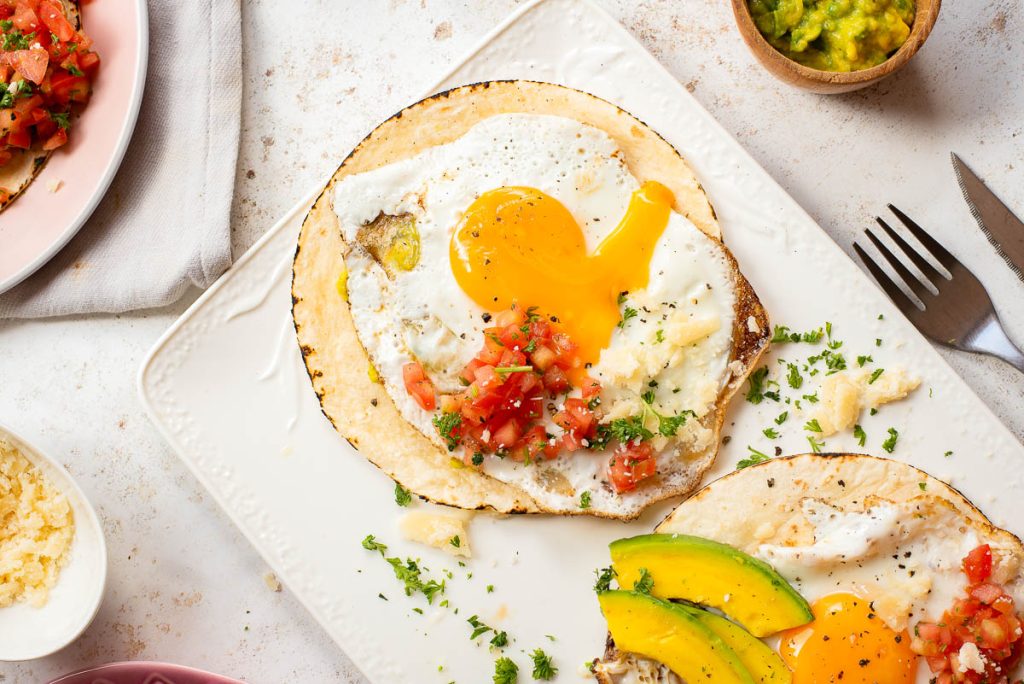 A plate with two corn tortillas topped with fried eggs, diced tomatoes, sliced avocado, chopped herbs, and cheese—this Fried Egg Taco dish is served with a fork and knife placed nearby.