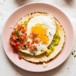 A Fried Egg Taco features a corn tortilla topped with guacamole, salsa, and a sunny-side-up egg, served on a pink plate.