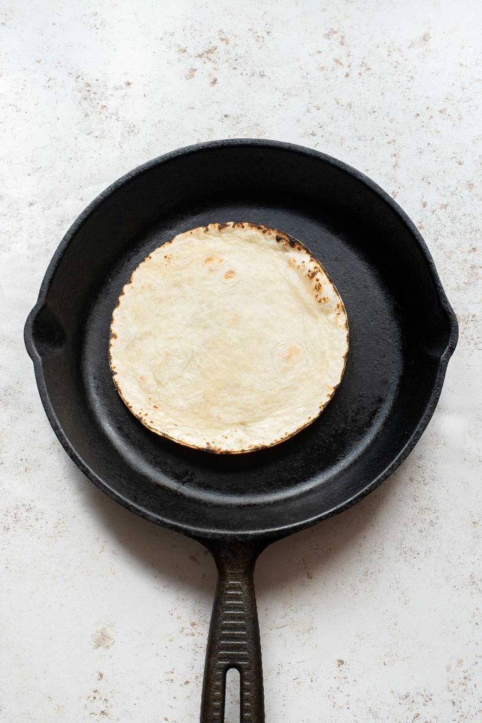 A single flour tortilla is heating in a black cast iron skillet on a light-colored surface, perfect for making a delicious Fried Egg Taco.