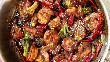 A pan filled with stir-fried shrimp, broccoli, dried red chilies, and sauce, garnished with sesame seeds and chopped green onions.