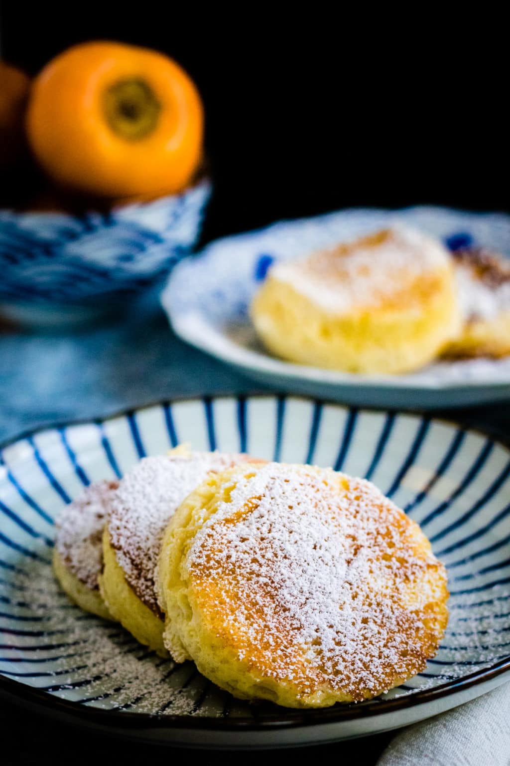 Three fluffy Japanese Souffle Pancakes dusted with powdered sugar rest on a striped plate, with more pancakes and fruit in the background.