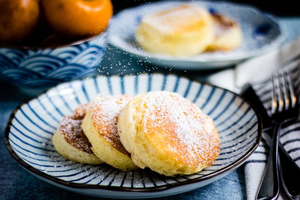 Three fluffy Japanese souffle pancakes on a striped plate are dusted with powdered sugar, with a bowl of fruit and another plate of pancakes in the background.