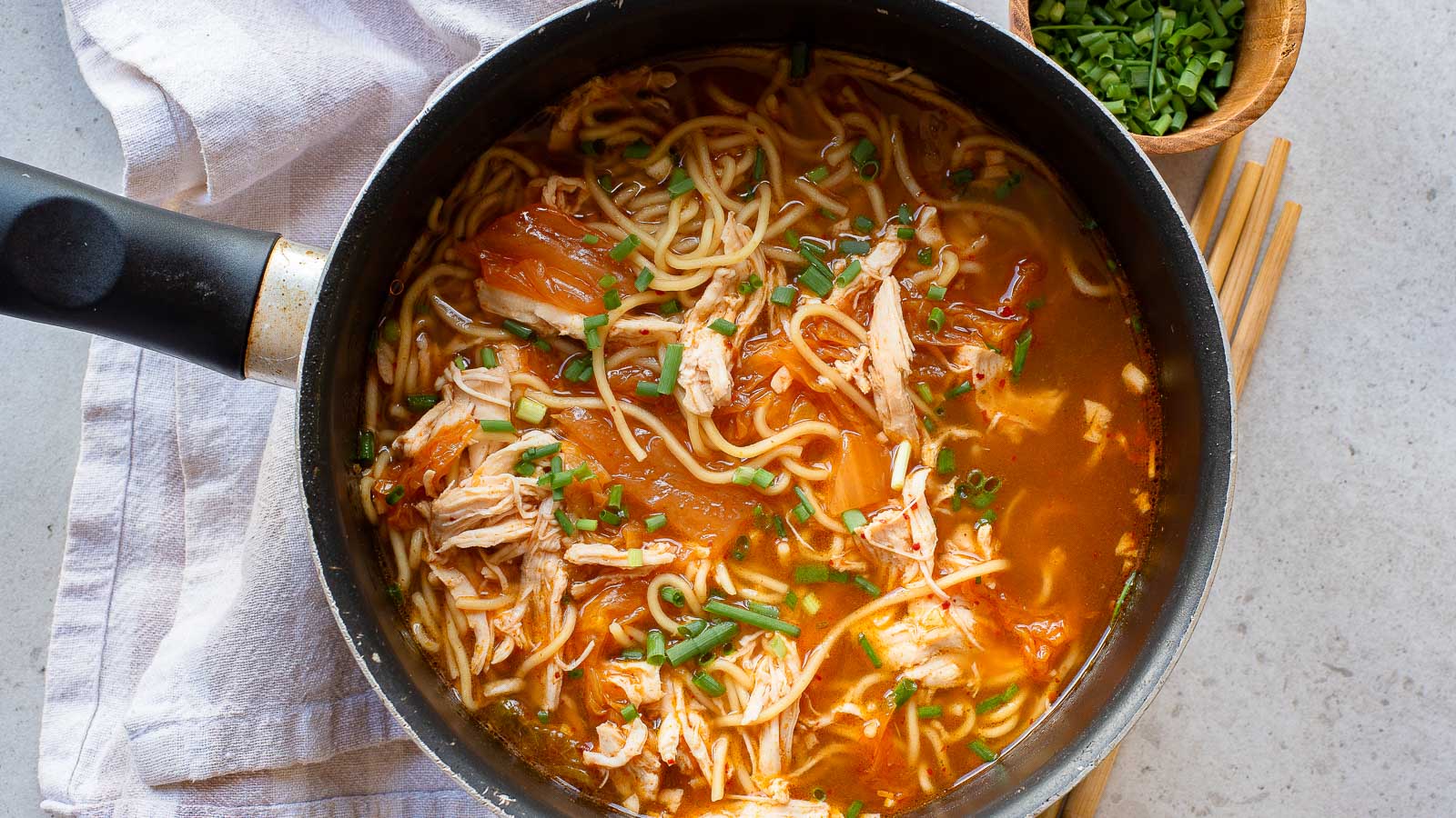 A pot of noodle soup with shredded chicken, green onions, and a red broth, placed on a white towel with wooden chopsticks and a bowl of chopped green onions nearby.