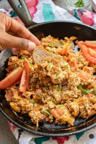 A hand dips a slice of bread into a skillet filled with Menemen—a tasty mix of Turkish scrambled eggs, tomatoes, and herbs; sliced tomatoes are arranged on the side.