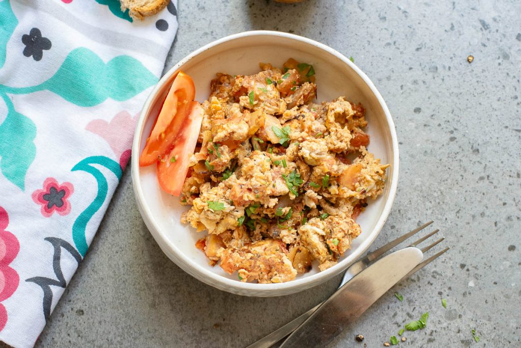 A white bowl filled with Menemen, Turkish scrambled eggs mixed with vegetables and herbs and garnished with tomato slices, sits on a gray surface beside a fork, knife, and a patterned cloth—a classic Turkish breakfast scene.