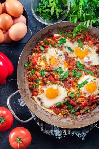 A skillet of Moroccan Shakshuka with poached eggs in a tomato and pepper sauce, garnished with parsley, surrounded by fresh eggs, tomatoes, a red pepper, and herbs—a delicious Moroccan breakfast classic.