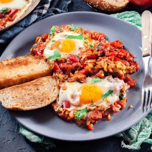 A gray plate with two eggs cooked in a flavorful tomato and vegetable sauce, inspired by a Moroccan Shakshuka recipe, served with toasted bread, a fork, and a knife.