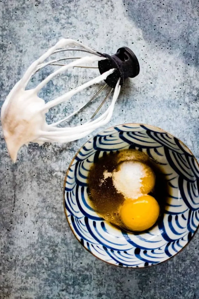 A patterned bowl with two raw eggs, sugar, and brown liquid sits next to a mixer whisk coated in whipped egg whites—essential ingredients for Japanese souffle pancakes—on a textured surface.