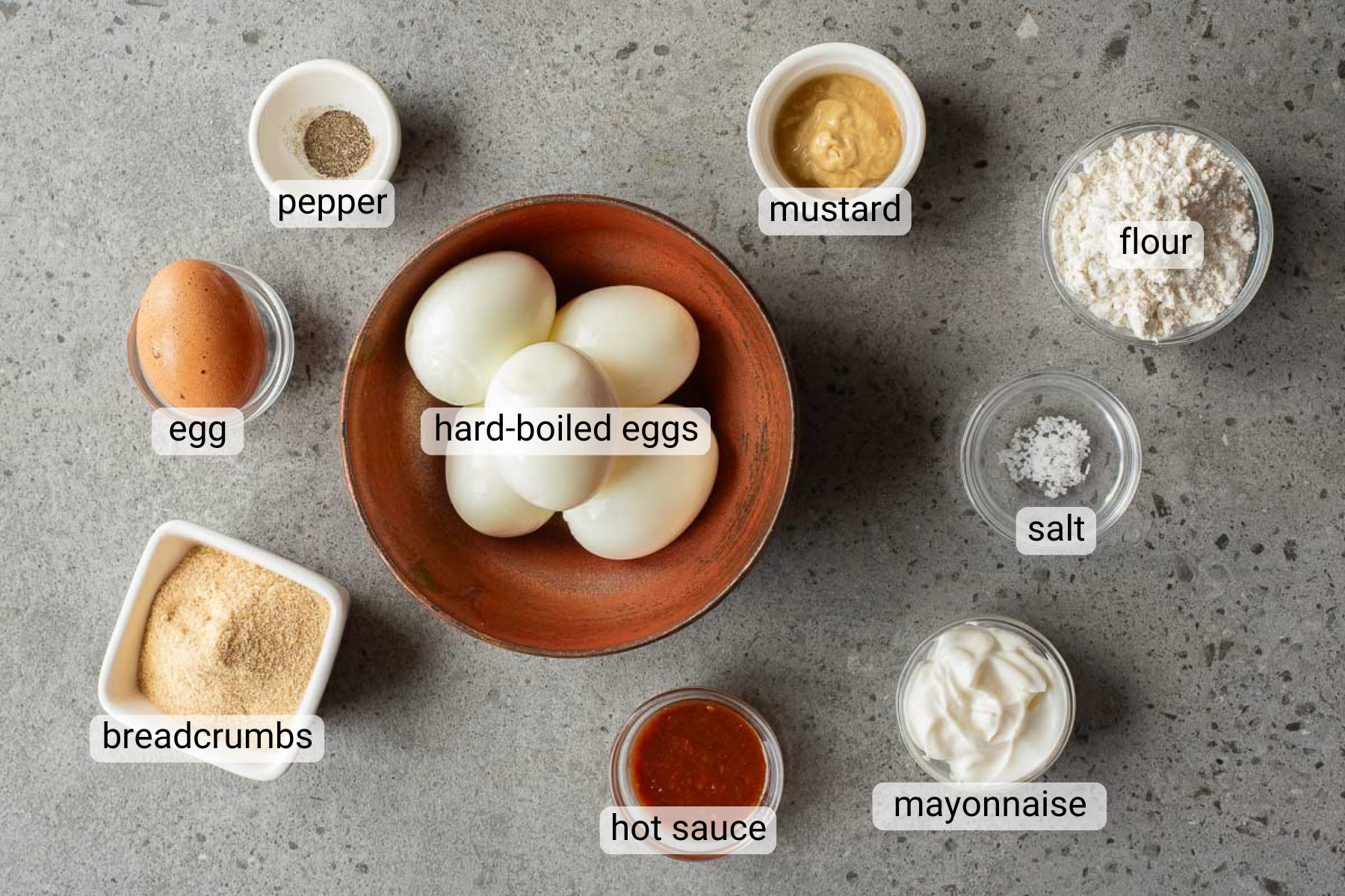 A bowl of hard-boiled eggs is surrounded by small bowls of pepper, mustard, flour, salt, mayonnaise, hot sauce, breadcrumbs, and a whole egg—perfect ingredients for making delicious Fried Deviled Eggs—arranged on a gray surface.