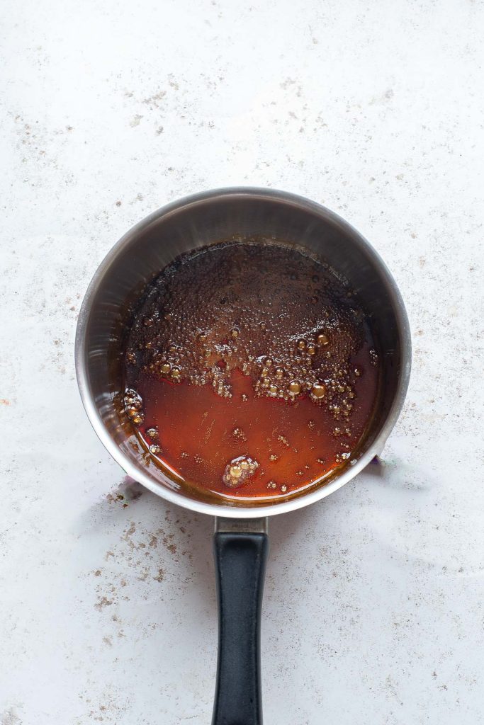 A stainless steel saucepan containing simmering coffee caramel with bubbles on the surface, placed on a light-colored countertop—perfect for making Vietnamese egg flan.