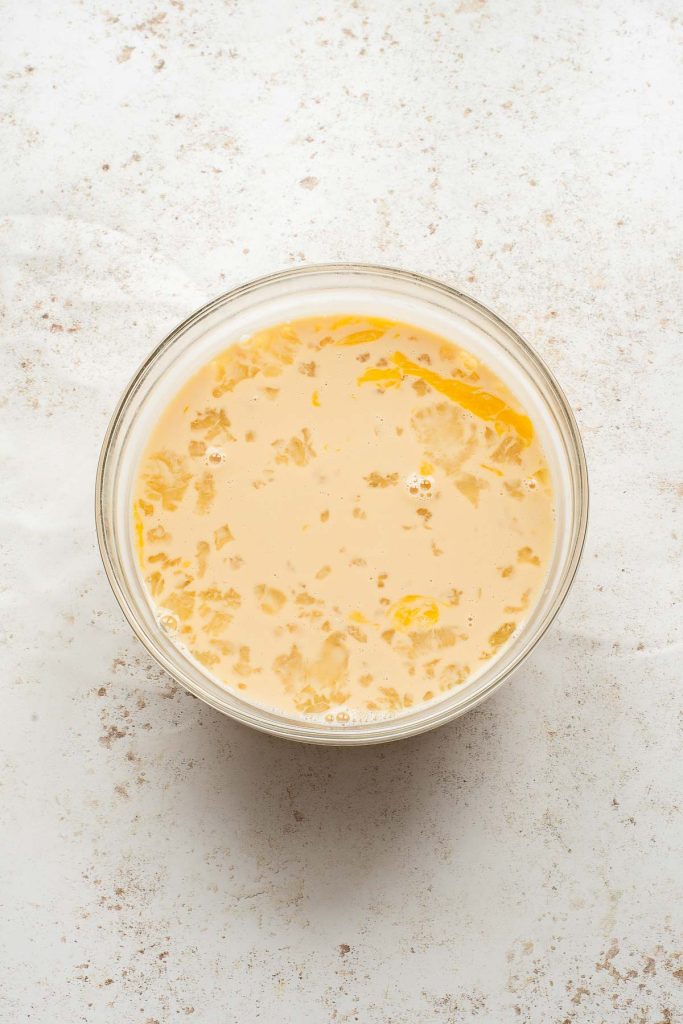 A glass bowl containing beige liquid with scattered yellow streaks, reminiscent of Vietnamese Egg Flan, set on a lightly speckled white surface.