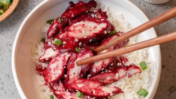 A bowl of white rice topped with sliced red-glazed meat, garnished with sesame seeds and chopped green onions, with chopsticks picking up a piece.