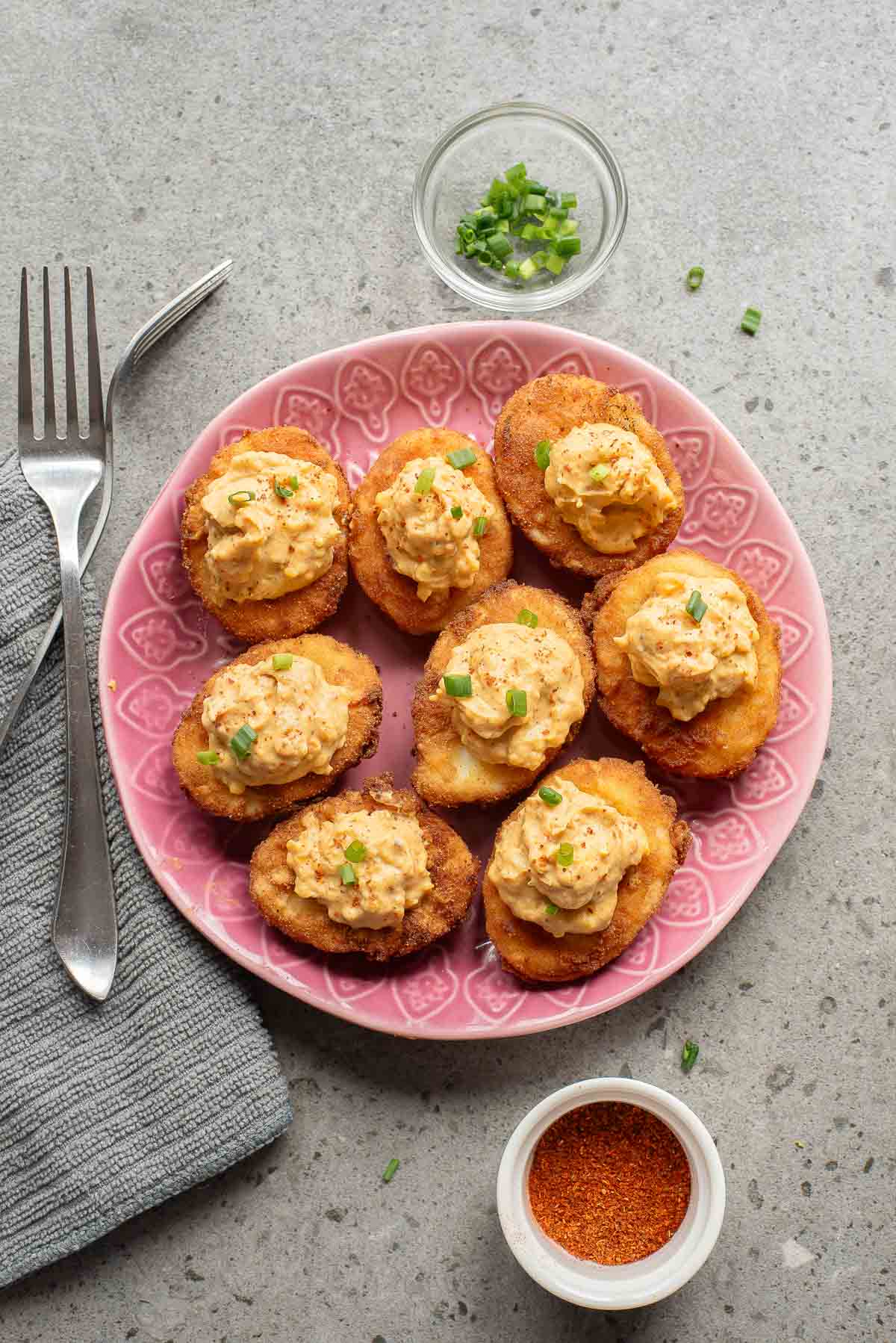 A pink plate with eight fried deviled eggs topped with a creamy mixture and chopped green onions, next to a fork, a bowl of green onions, and a bowl of spice on a gray surface.