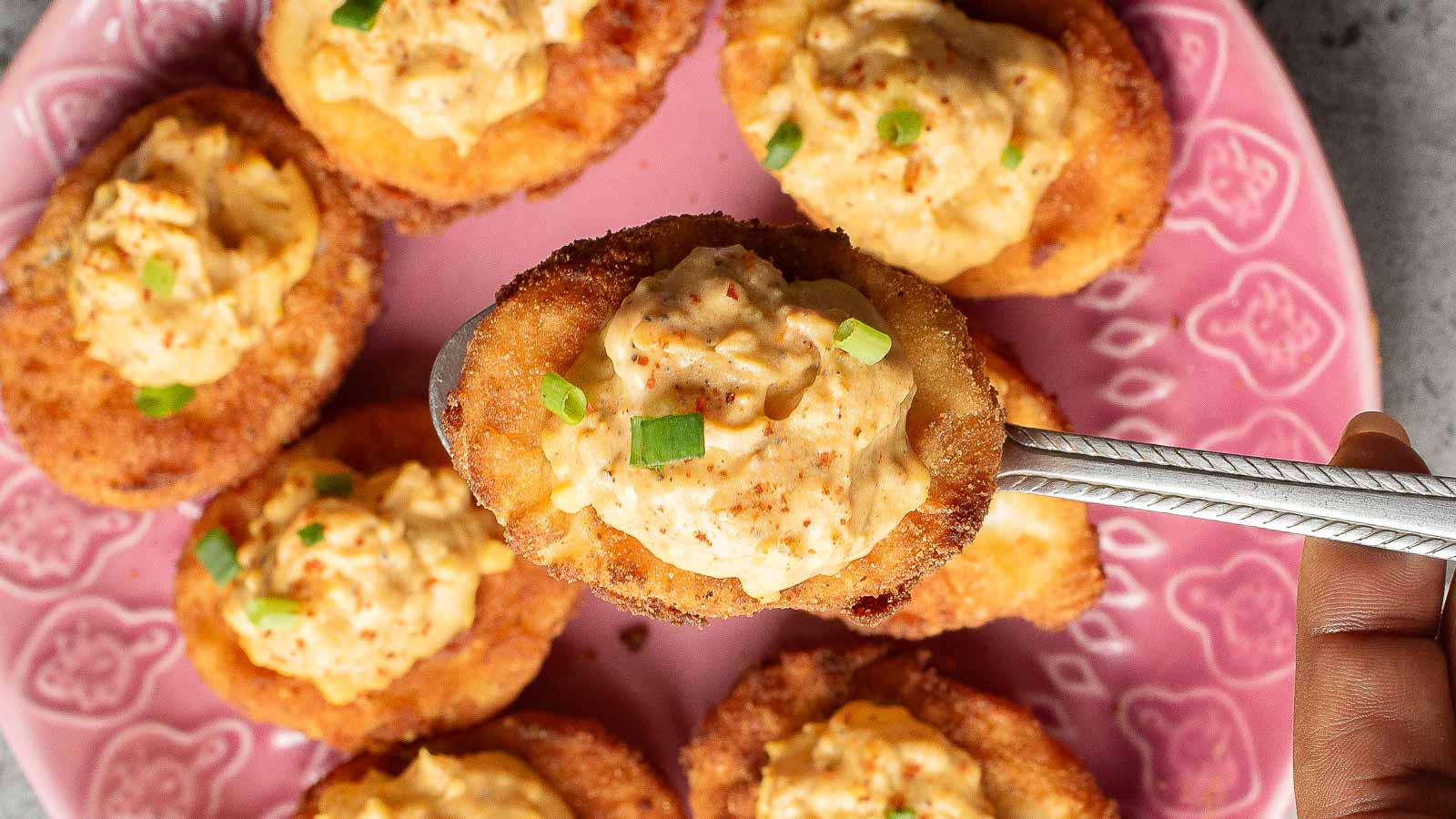 A hand holds a spoon with a breaded, fried deviled egg topped with creamy, orange filling and chopped green onions; several more fried deviled eggs rest on a pink plate below.