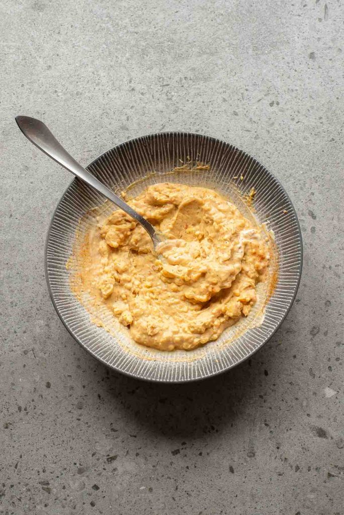 A bowl of creamy, orange-colored deviled eggs mixture with a spoon, set on a gray speckled surface.