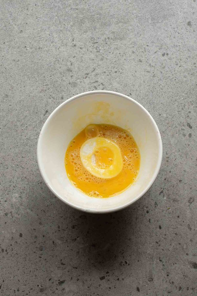 A white bowl containing a lightly beaten egg mixture for Fried Deviled Eggs sits on a gray speckled countertop.