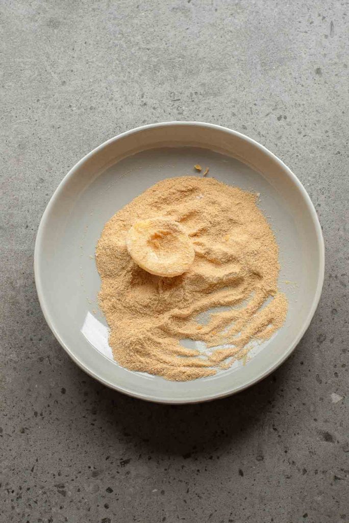 A white plate with a mound of yellow powder and a round food item, resembling Fried Deviled Eggs, partially coated in the powder, on a gray speckled surface.