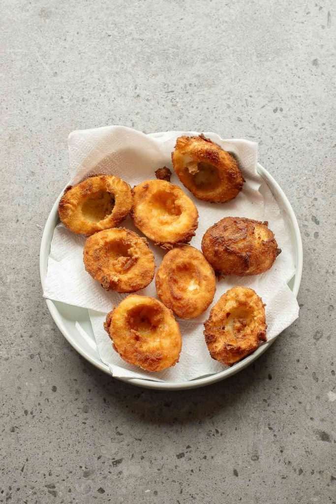 A plate with eight golden-brown fried deviled eggs rests on paper towels, set on a gray speckled surface.