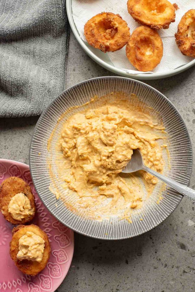 A bowl of egg salad with a spoon, fried deviled eggs rings drying on a paper towel, and some topped with egg salad on a pink plate nearby.