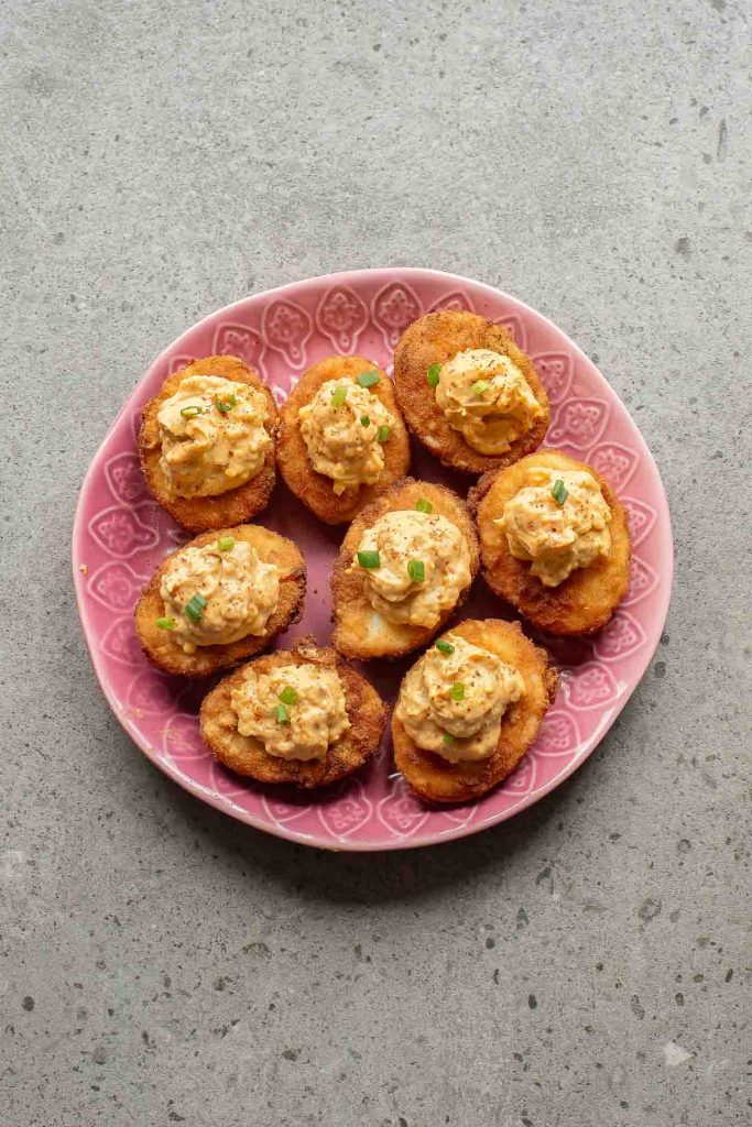 A pink plate with eight pieces of fried bread topped with a creamy fried deviled eggs mixture and garnished with chopped herbs on a gray surface.