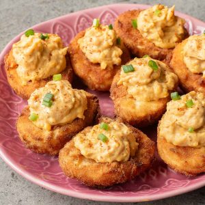 A pink plate with eight crispy fried deviled eggs, topped with a creamy filling and garnished with chopped green onions—a delicious twist on the classic deviled eggs recipe.
