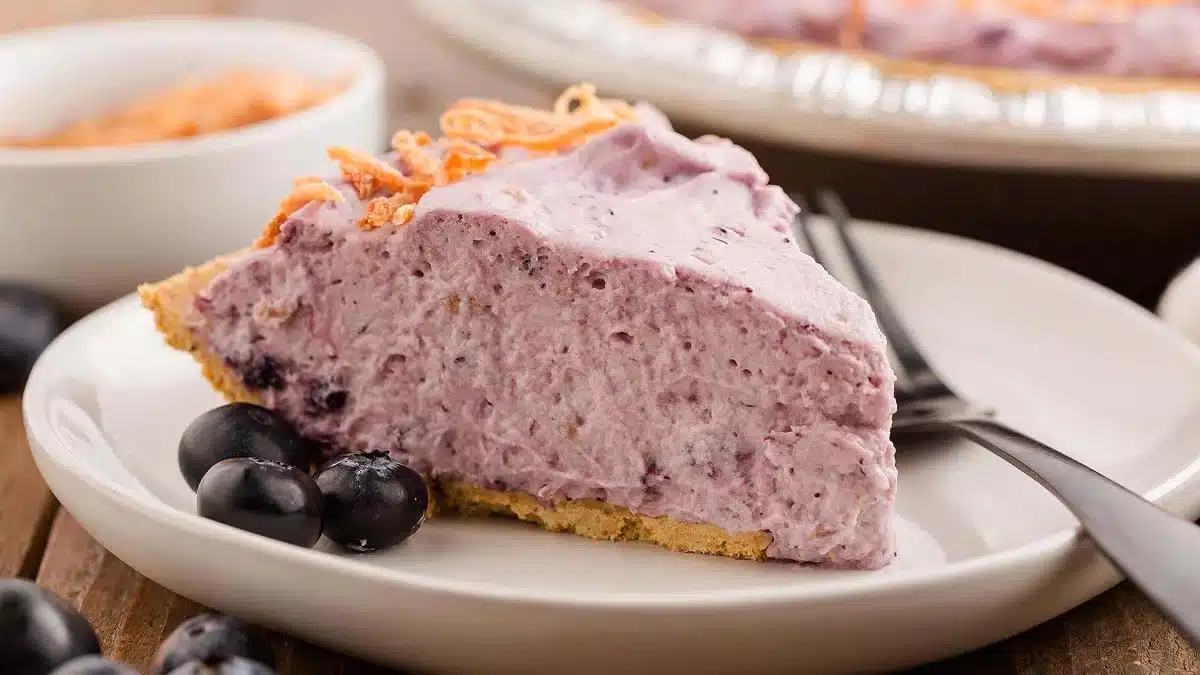 A slice of blueberry cream pie sits on a white plate with a fork, garnished with fresh blueberries.