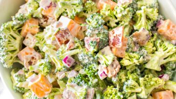 A bowl of broccoli salad with cheddar cheese cubes, chopped red onion, bacon bits, and creamy dressing—perfect for those who love Classic Recipes.