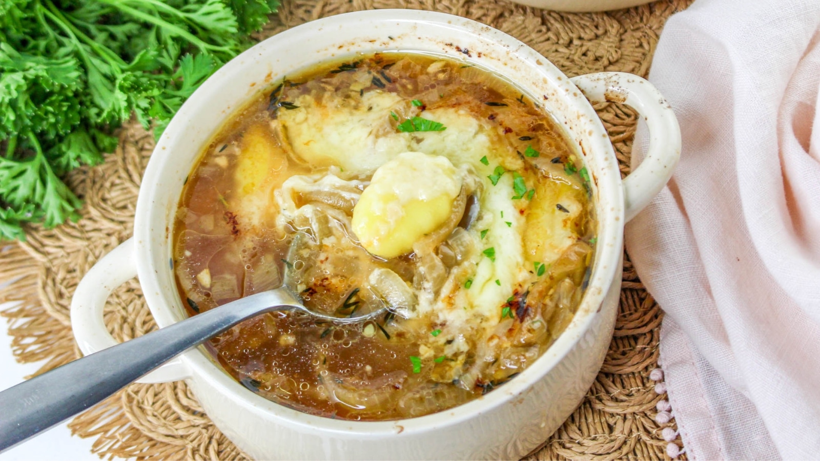 A bowl of onion soup topped with melted cheese and herbs, with a spoon resting inside. Fresh parsley is visible in the background.