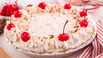 A cream pie with a whipped topping, chopped nuts, and maraschino cherries, served in a foil pie tin on a table with striped napkins—just like one of Grandma’s Kitchen classic recipes.