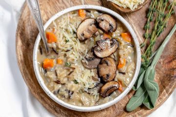 A bowl of creamy wild rice soup with sautéed mushrooms, diced carrots, shredded cheese, and fresh herbs, served on a wooden plate with a spoon.