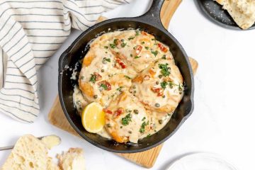 Cast iron skillet with chicken breasts in a creamy sauce—an ultimate comfort food classic, garnished with parsley, capers, sun-dried tomatoes, and a lemon wedge; bread and striped towel nearby.