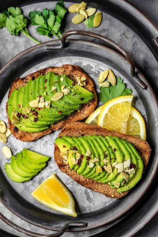 Two slices of toast topped with sliced avocado, nuts, and spices on a metal tray, with lemon wedges and parsley garnish—perfect for using avocados after freezing avocados to enjoy later.