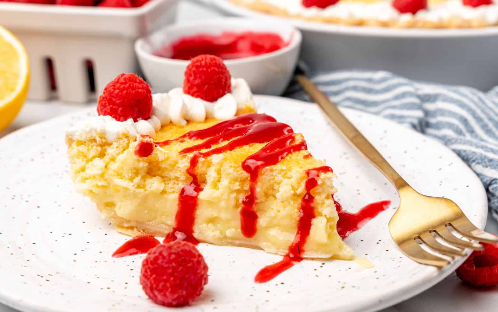 A slice of custard pie topped with whipped cream, fresh raspberries, and raspberry sauce on a white plate with a gold fork.