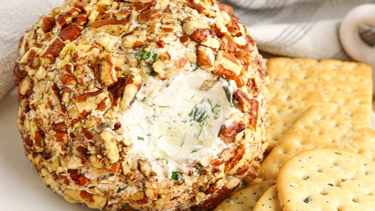 A garlic herb cheese ball coated with chopped nuts sits on a plate, surrounded by a variety of crackers. A portion of the cheeseball has been scooped out, revealing its creamy interior. A striped cloth is visible in the background