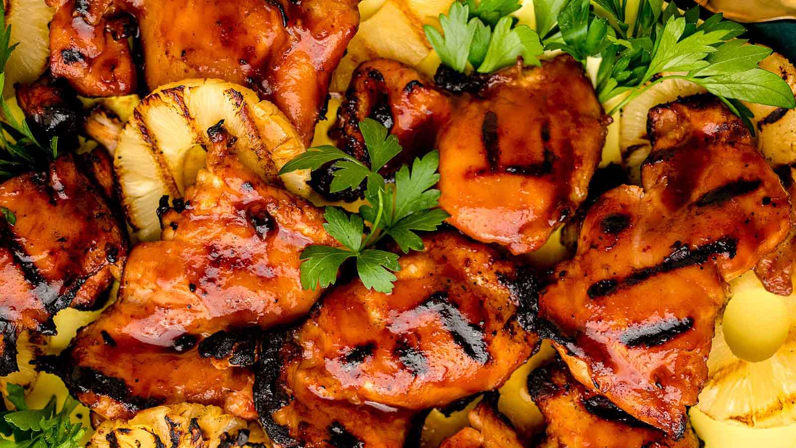 Grilled pieces of glazed chicken are arranged on a plate with slices of grilled pineapple and garnished with fresh parsley.