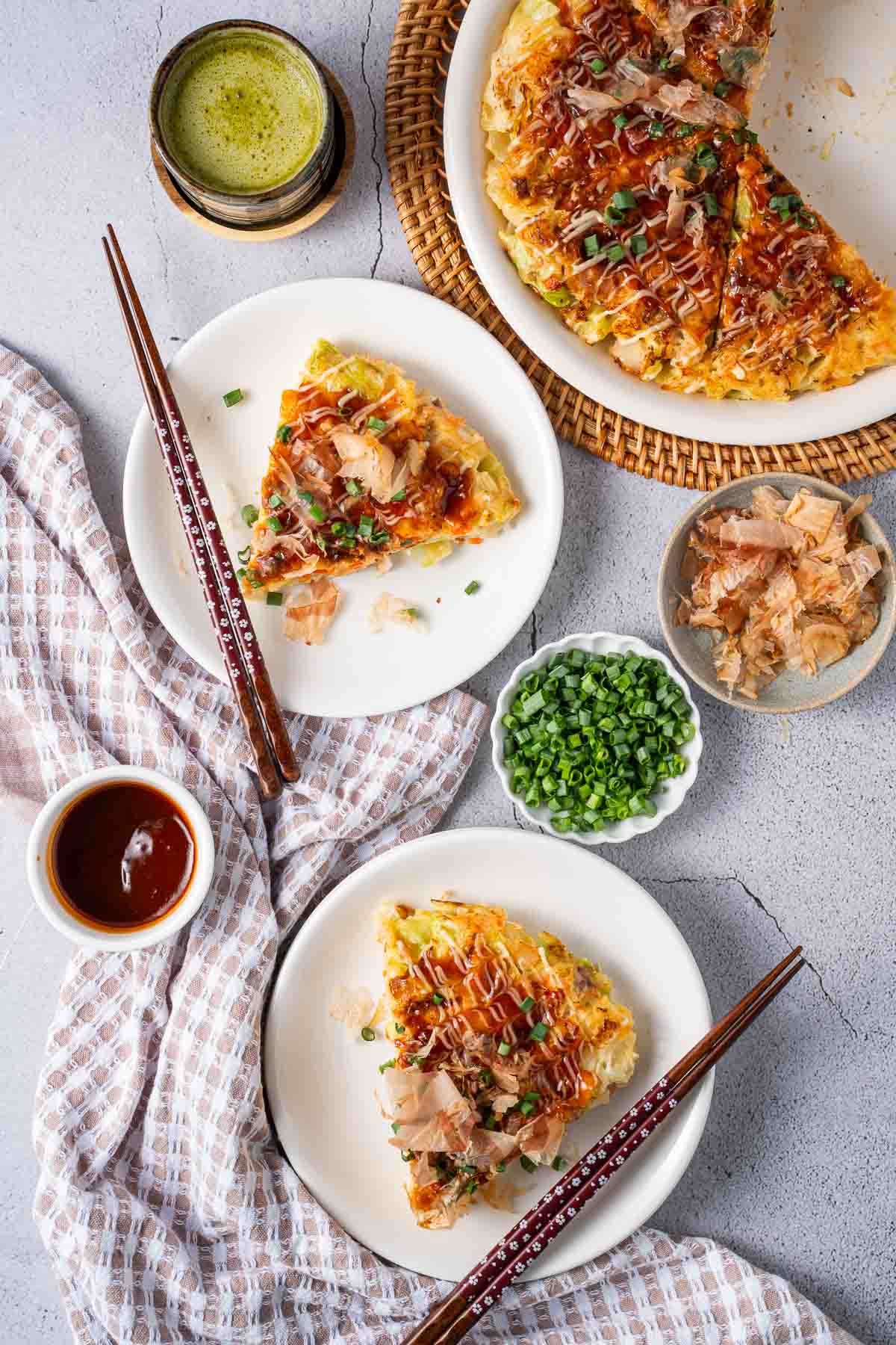 Two plates with slices of okonomiyaki, topped with green onions and bonito flakes, are set on a table with chopsticks, sauce, extra toppings, and a drink nearby.
