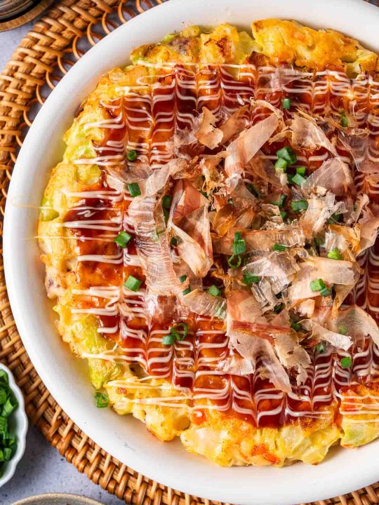 A plate of okonomiyaki topped with mayonnaise, okonomiyaki sauce, bonito flakes, and chopped green onions, served on a woven placemat.