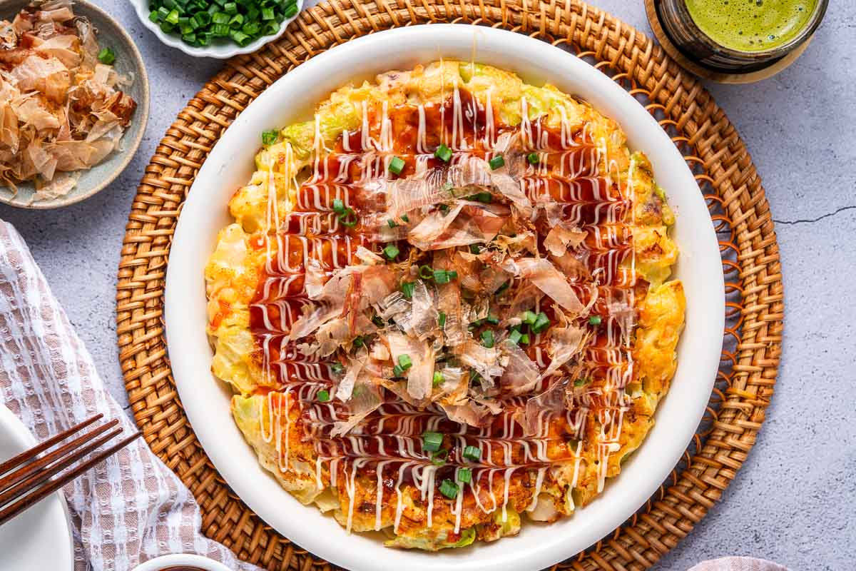 A plate of okonomiyaki, a Japanese savory pancake, topped with sauce, mayonnaise, bonito flakes, and chopped green onions, on a woven placemat.